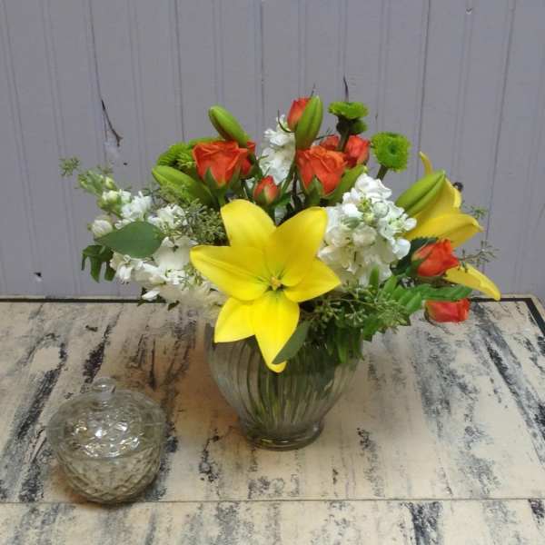 Bouquet of yellow lilies, orange roses, and white flowers in a glass vase