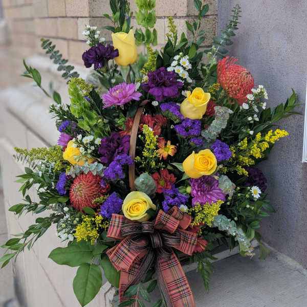 Basket arrangement with yellow roses, purple blooms, and a plaid ribbon