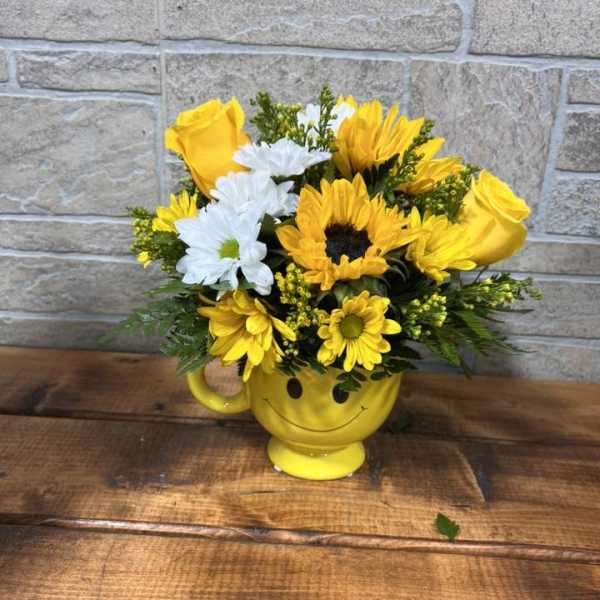 Yellow and white flowers arranged in a yellow smiley-face mug on a wooden surface.