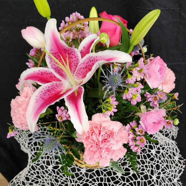 Pink lilies and carnations arranged in a basket with small purple flowers.