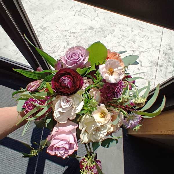 Handheld bouquet of roses, ranunculus, and pale blooms in pink, burgundy, and cream