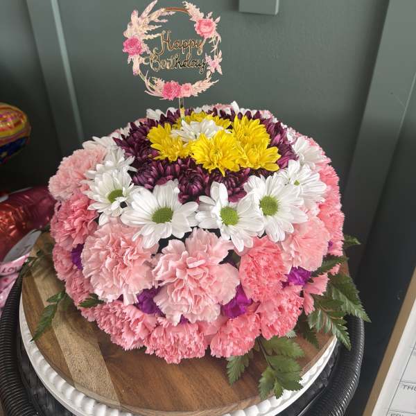 Round floral cake of pink carnations and mixed daisies with Happy Birthday topper on a wooden stand