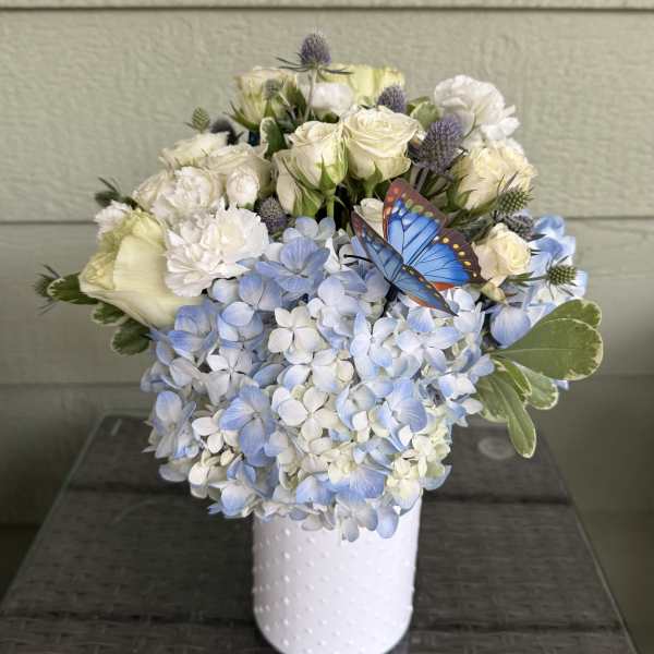 White roses and blue hydrangeas in a white vase with a butterfly accent