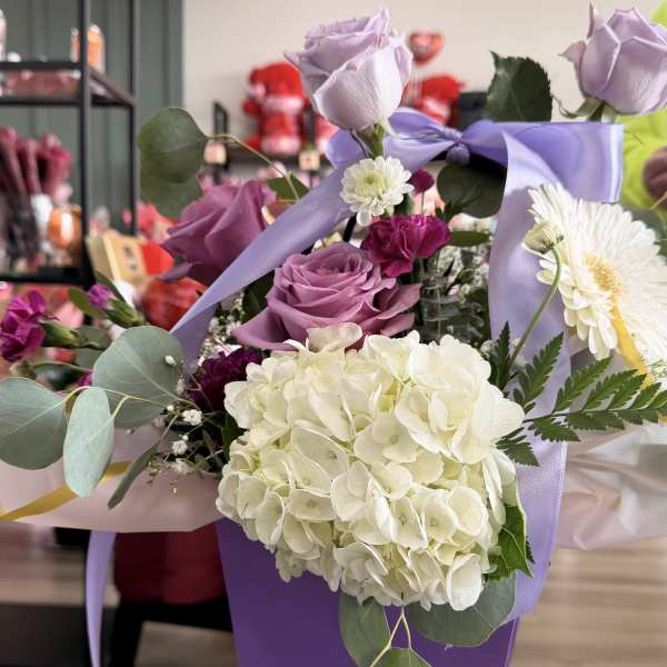 Purple box arrangement with lavender roses, white hydrangea, and mixed blooms accented by a satin bow.