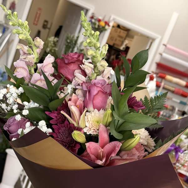 Hand-tied bouquet of pink roses, lilies, and mixed blooms wrapped in burgundy paper with a gold ribbon.