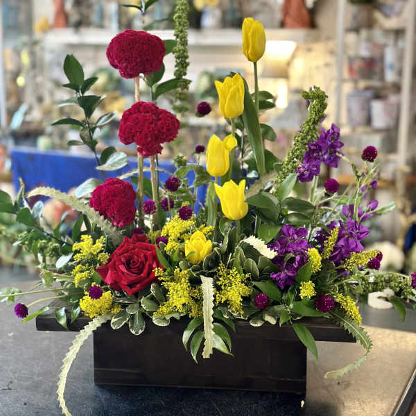 Mixed floral arrangement with red roses, yellow tulips, and purple blooms in a black container