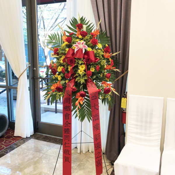 Tall standing floral spray with red ribbons and mixed red, yellow, and orange flowers