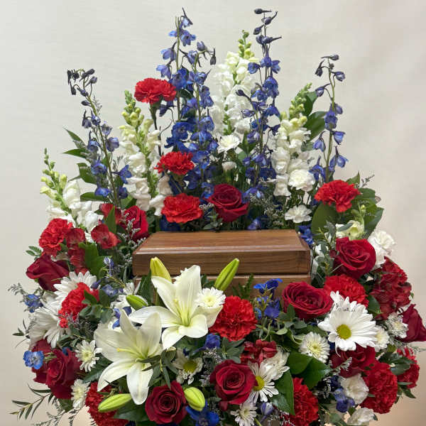 Large funeral spray with red roses, white lilies, and blue delphinium around a wooden urn