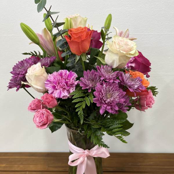 Mixed bouquet of roses, lilies, and purple mums in a glass vase with a pink ribbon