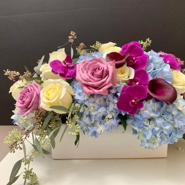 Arrangement of pink and cream roses with blue hydrangeas and purple orchids in a white container
