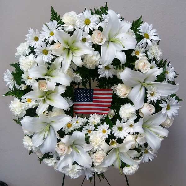 White floral wreath with lilies, daisies, and roses around an American flag