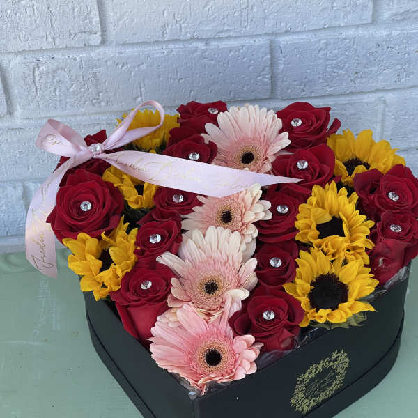 Heart-shaped box of red roses, pink gerbera daisies, and yellow sunflowers with a ribbon