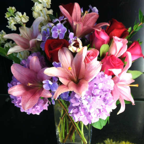 Pink lilies, red and pink roses, and purple hydrangeas in a clear glass vase