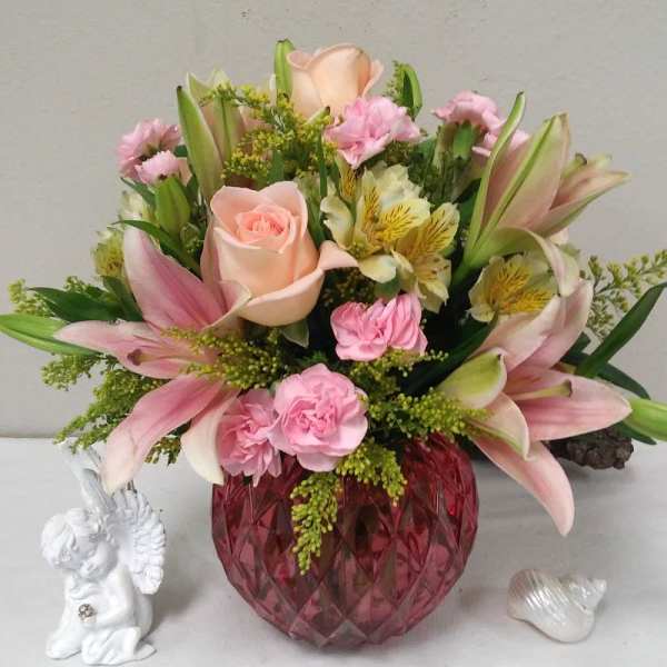 Pink lilies and roses arranged in a red glass vase with a small angel figurine