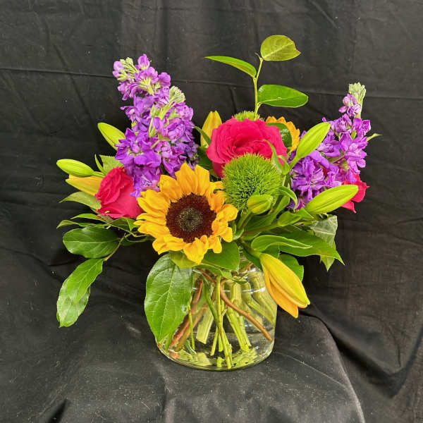 Mixed bouquet in a clear glass vase with sunflowers, pink roses, and purple blooms