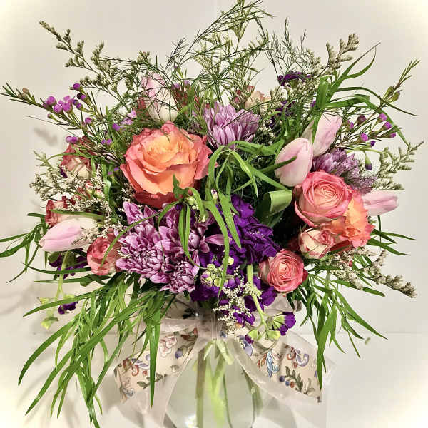 Mixed bouquet of pink roses, purple mums, and tulips with trailing greenery in a glass vase