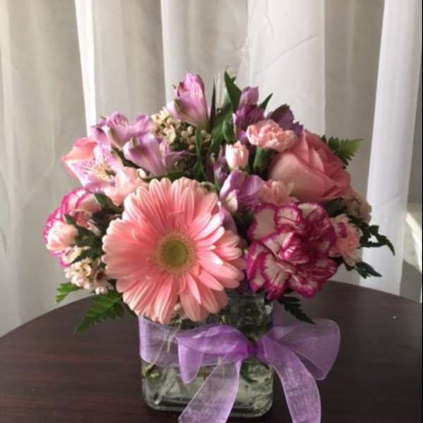 Compact pink and purple flower arrangement in a clear glass cube vase with a lavender ribbon bow