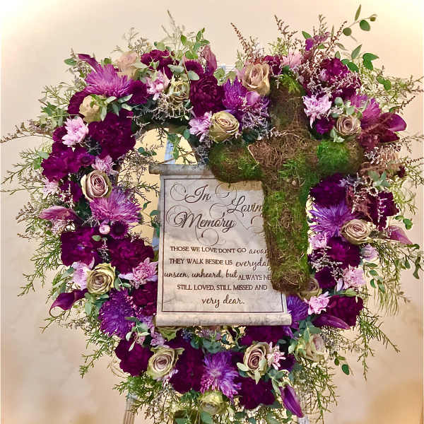 Standing sympathy wreath of purple and cream flowers with a moss-covered cross and memorial plaque on an easel