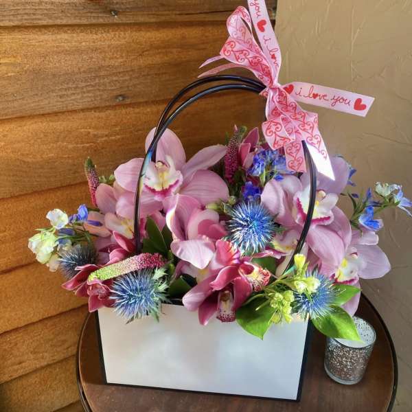 Pink orchid arrangement in a white gift bag with a heart ribbon