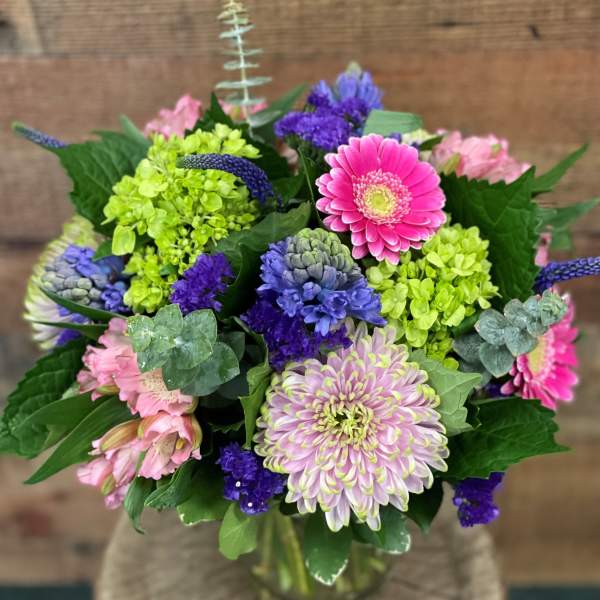 Colorful bouquet with pink gerbera daisies, green hydrangeas, and purple blooms in a glass vase