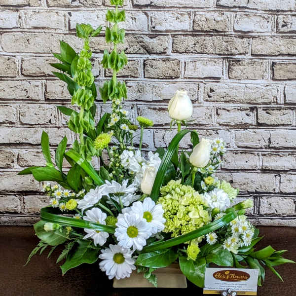 Rectangular arrangement of white daisies, white roses, green hydrangea, and tall green Bells of Ireland.
