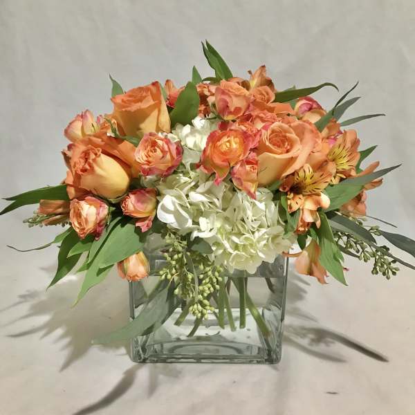 Short arrangement of orange roses and white hydrangeas in a clear glass cube vase