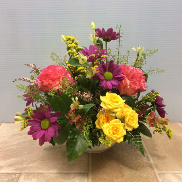 Mixed bouquet of yellow roses, purple daisies, and pink carnations in a white bowl vase