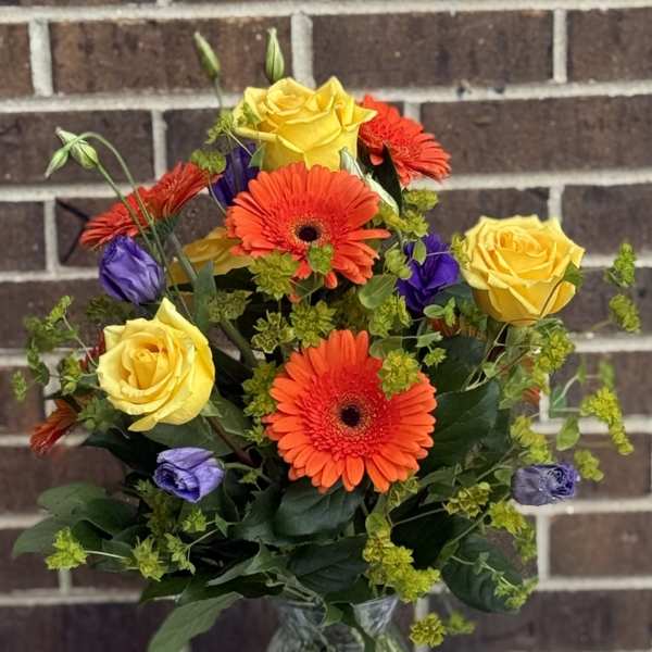 Bouquet of yellow roses, orange gerbera daisies, and purple blooms in a glass vase