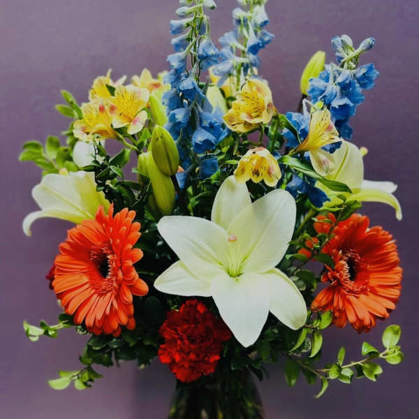 Mixed bouquet with white lilies, orange gerbera daisies, blue flowers, and red carnations in a vase