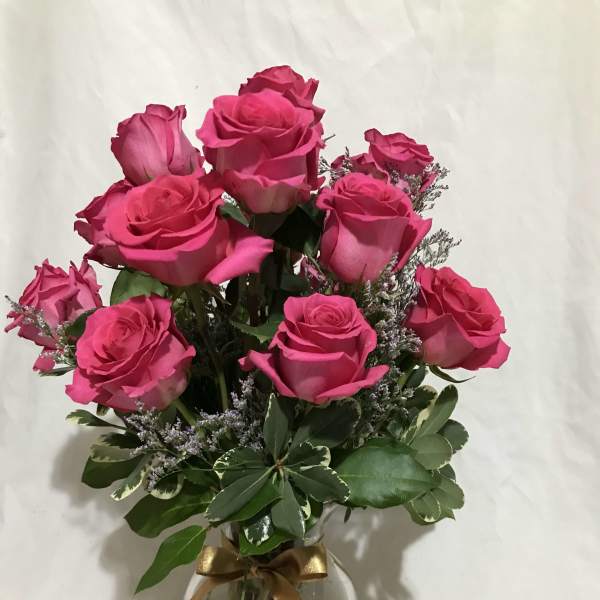 Arrangement of bright pink roses with filler blooms in a clear glass vase tied with a gold ribbon.