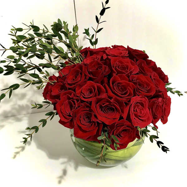 Round arrangement of red roses in a clear glass bowl vase with trailing greenery accents.