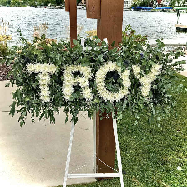 Standing tribute spray spelling TROY in white flowers with lush greenery on an easel