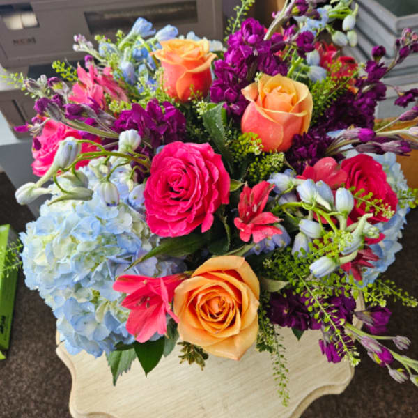 Bright bouquet with orange and pink roses, blue hydrangeas, and purple flowers on a small pedestal table
