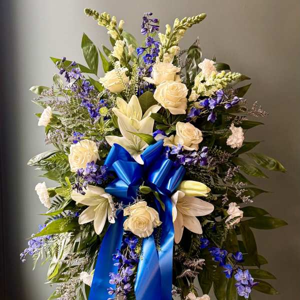Standing spray of white roses and lilies with blue flowers and a large blue bow on an easel.