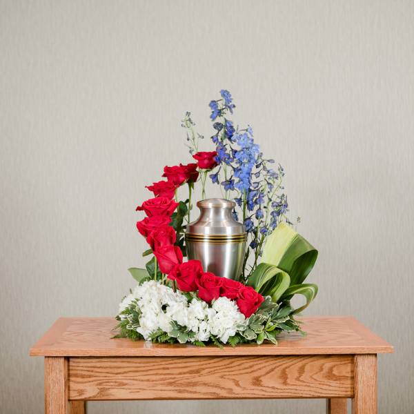 Red roses and blue flowers arranged around a silver urn on a table.
