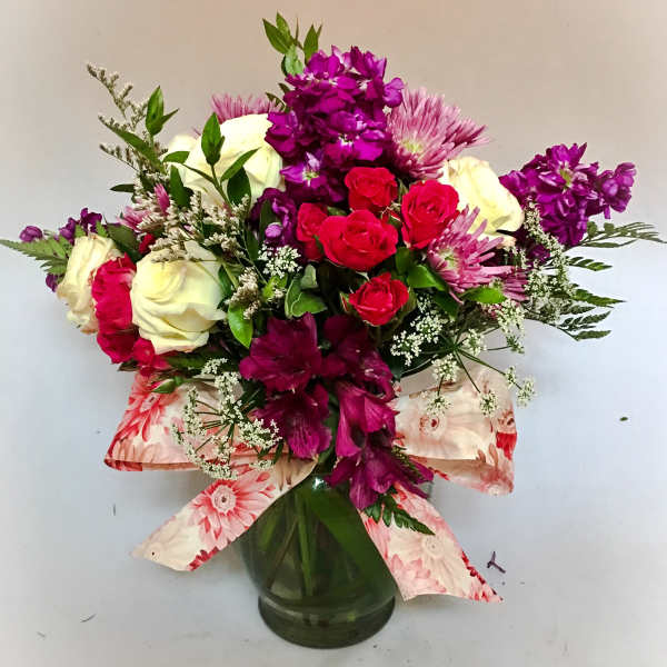 Mixed arrangement of white roses, red spray roses, and purple flowers in a glass vase with a floral ribbon bow