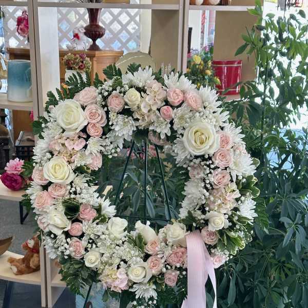 Heart-shaped wreath of white and blush flowers on a stand with a pink ribbon