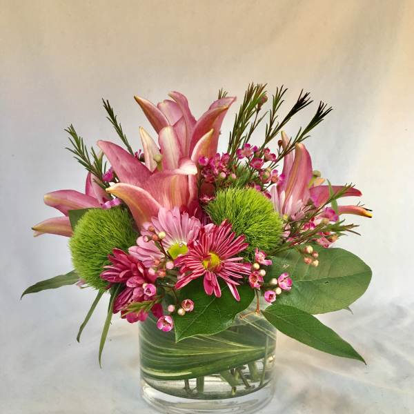 Compact arrangement of pink lilies and daisies with green pom blooms in a leaf-wrapped glass vase