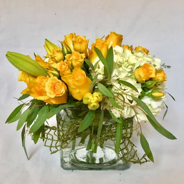 Low arrangement of yellow roses, lily buds, and white hydrangea in a clear glass cube vase