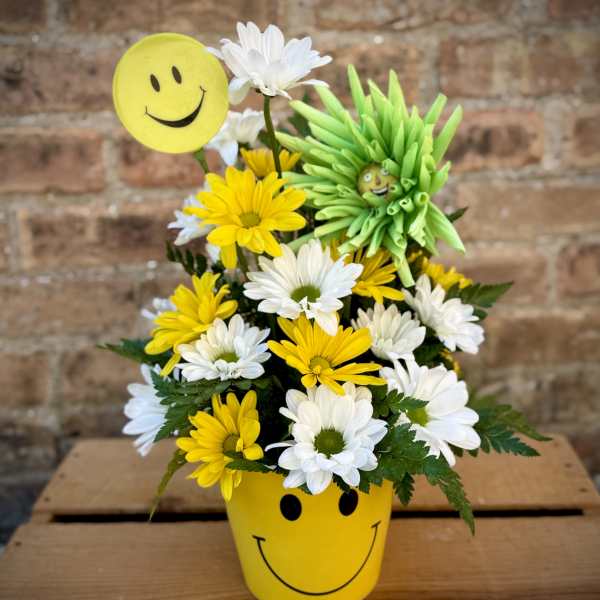 Yellow smiley-face pot holding white and yellow daisies with a smiley pick