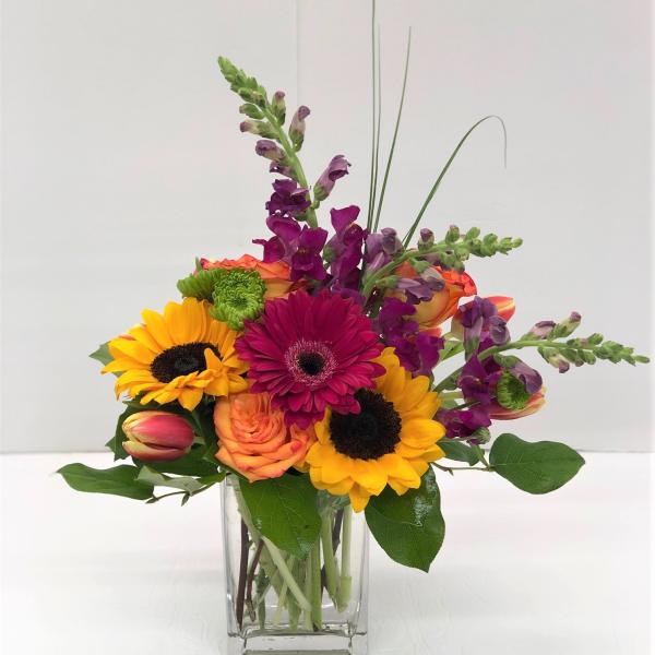 Mixed bouquet in a clear glass vase with sunflowers, roses, and purple spikes