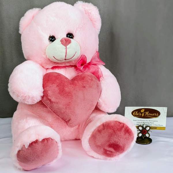 Pink teddy bear plush holding a darker pink heart pillow with a bow at its neck on a neutral backdrop
