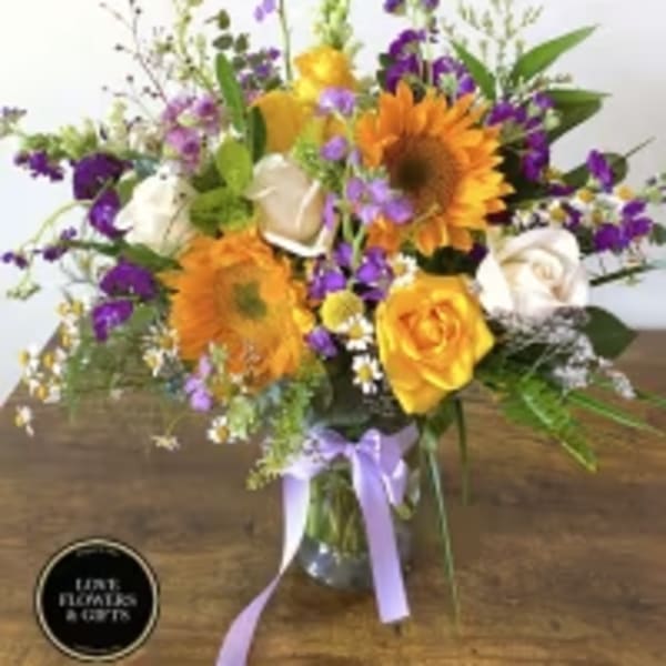 Mixed arrangement of yellow sunflowers, roses, and purple flowers in a clear vase with a lavender bow