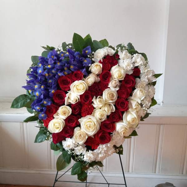 Heart-shaped floral tribute with red, white, and blue flowers on a stand