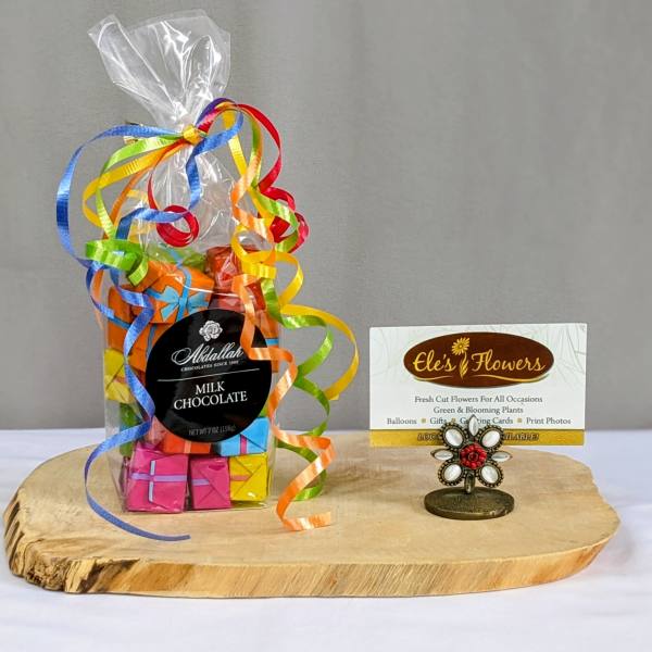 Bag of colorful wrapped milk chocolates with curling ribbons on a wooden board beside a florist business card.
