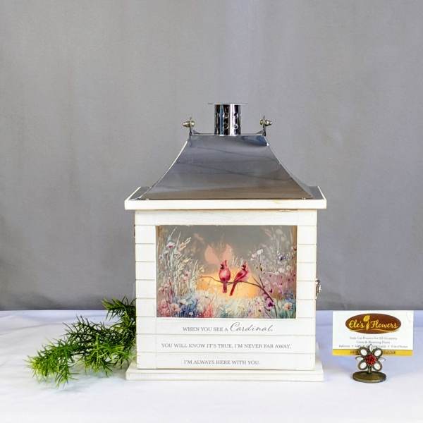 White lantern with cardinal artwork and a small greenery sprig beside it