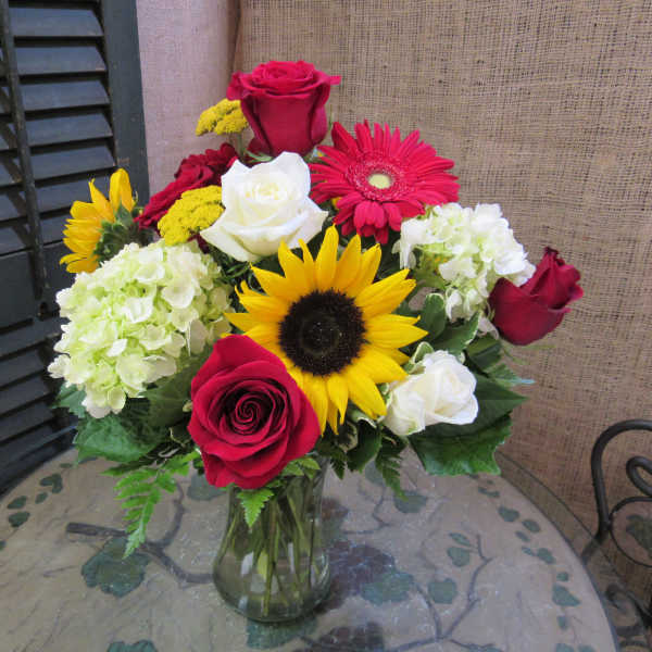 Mixed bouquet of roses, sunflowers, gerbera daisies, and hydrangeas in a glass vase