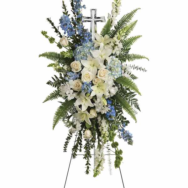 Standing floral spray with white lilies, cream roses, and blue flowers on a cross easel