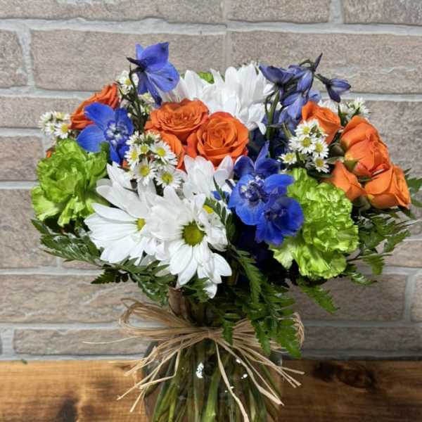Mixed bouquet of orange roses, blue blooms, white daisies and green carnations in a glass vase with raffia bow