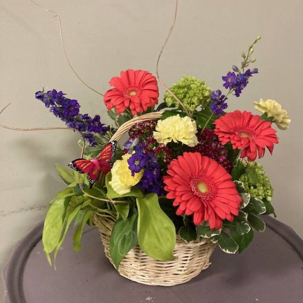 Basket arrangement with pink gerbera daisies, purple flowers, and yellow carnations